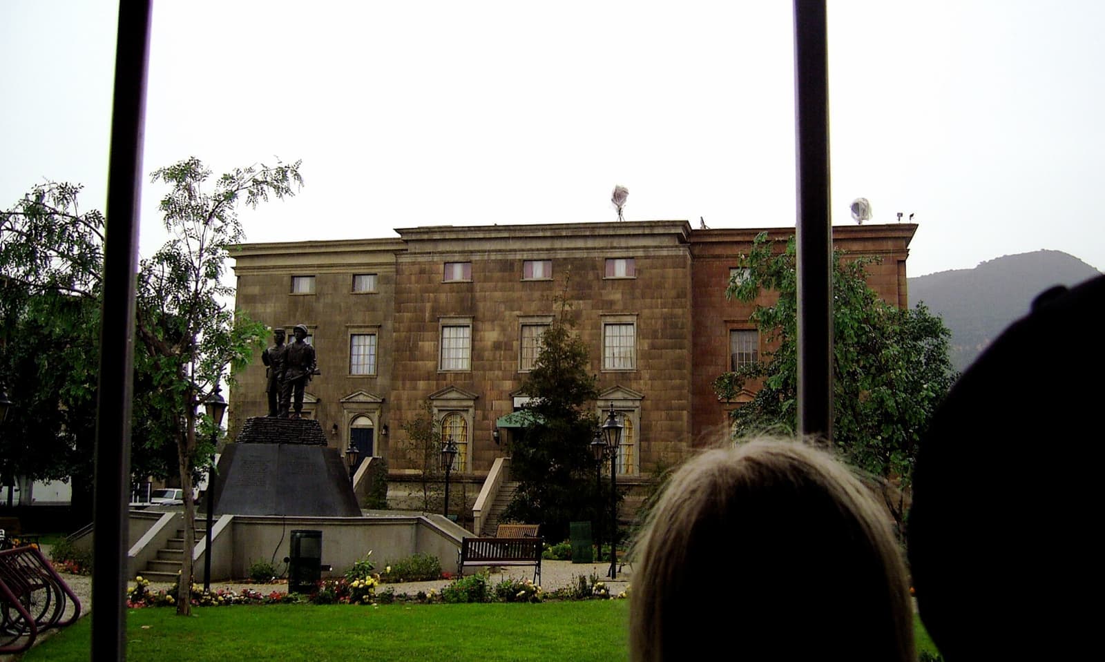 A pale classical courthouse with a clock tower behind a lawn, seen past the heads of tram passengers