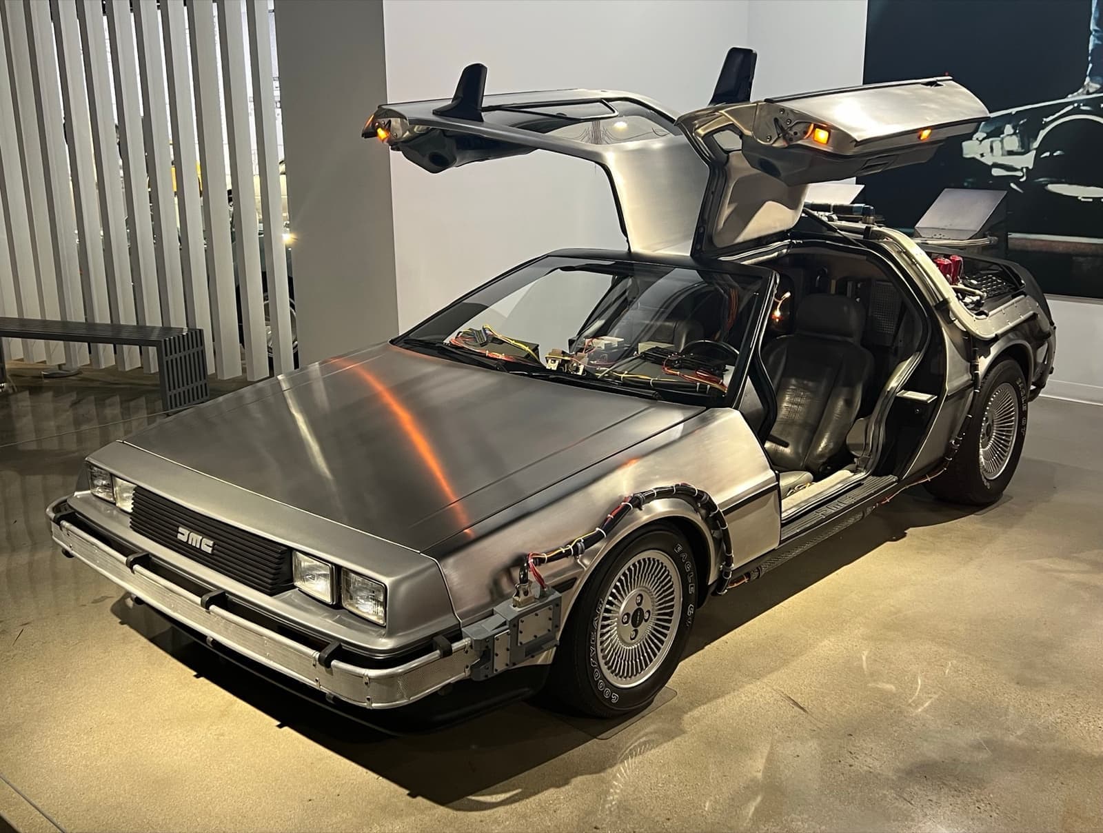 A stainless-steel DeLorean with gull-wing doors open, fitted with cables and vents, in a white gallery