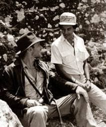 Harrison Ford in a fedora and leather jacket sitting on rocks beside a man in a cap, black and white