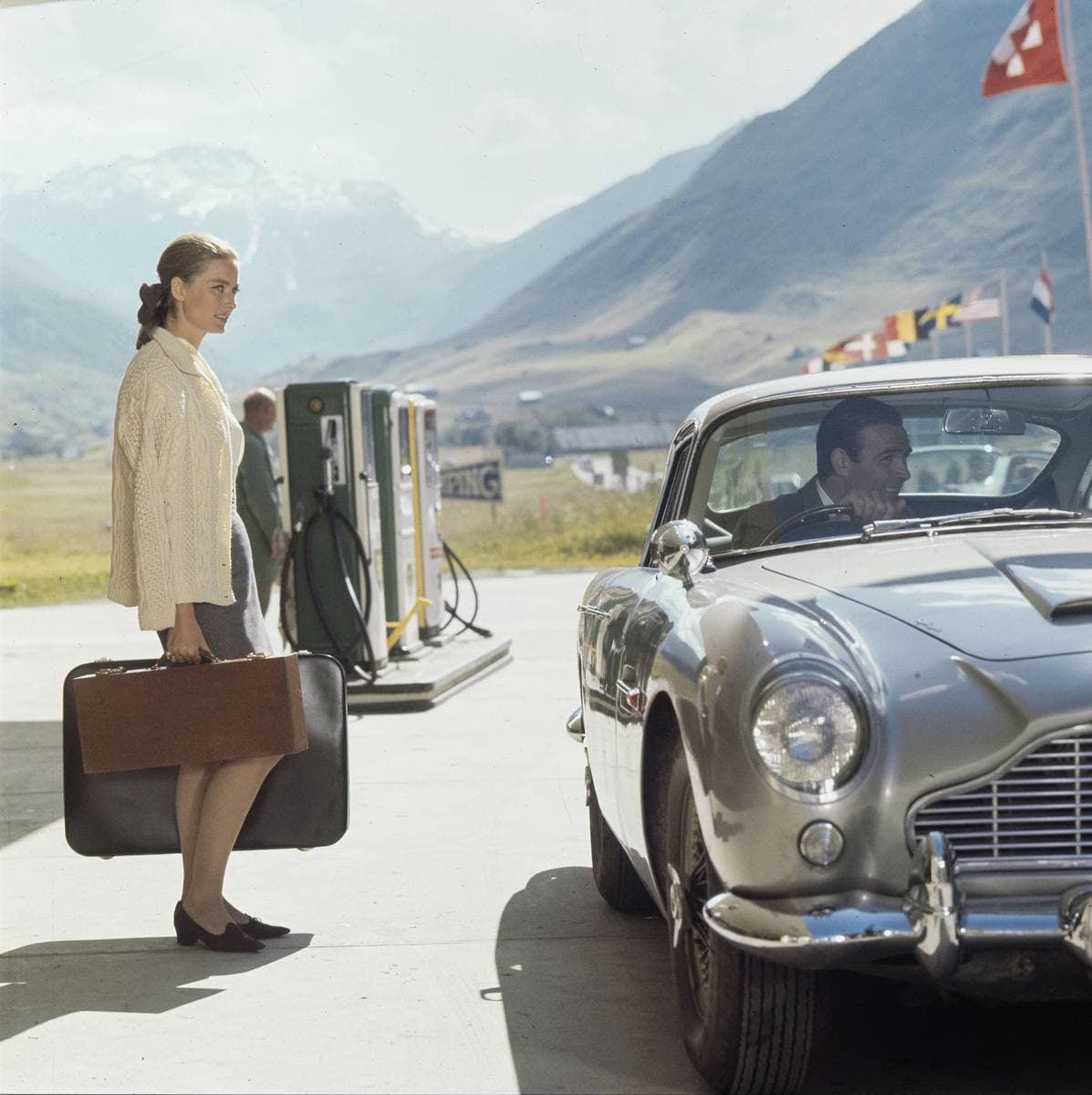 A woman with a suitcase beside petrol pumps and a silver sports car, Swiss mountains behind
