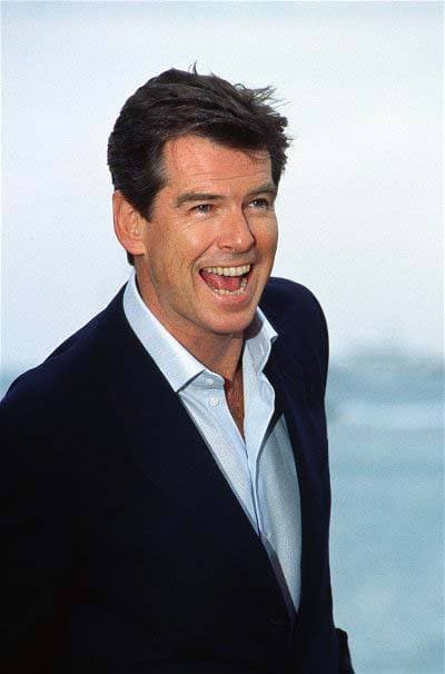 Pierce Brosnan laughing, in a navy jacket, sea behind