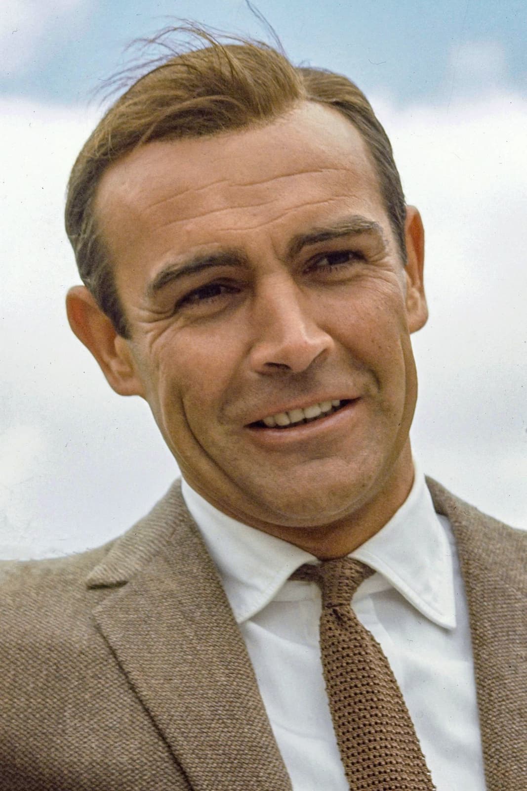 Sean Connery in a tweed jacket and tie, smiling, against a pale sky