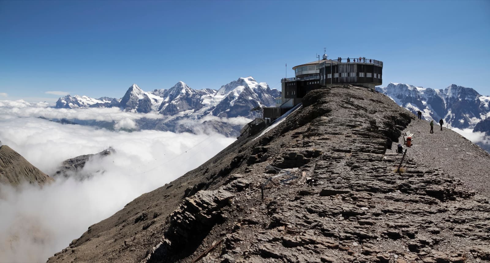 A rocky ridge rising out of cloud to a round summit building, with snow peaks behind