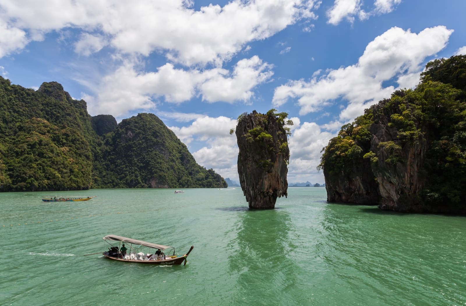 A narrow limestone spire standing in green water, a long-tail boat passing