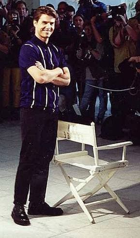 Tom Cruise in a dark short-sleeved shirt, arms folded, standing beside a director’s chair in front of photographers