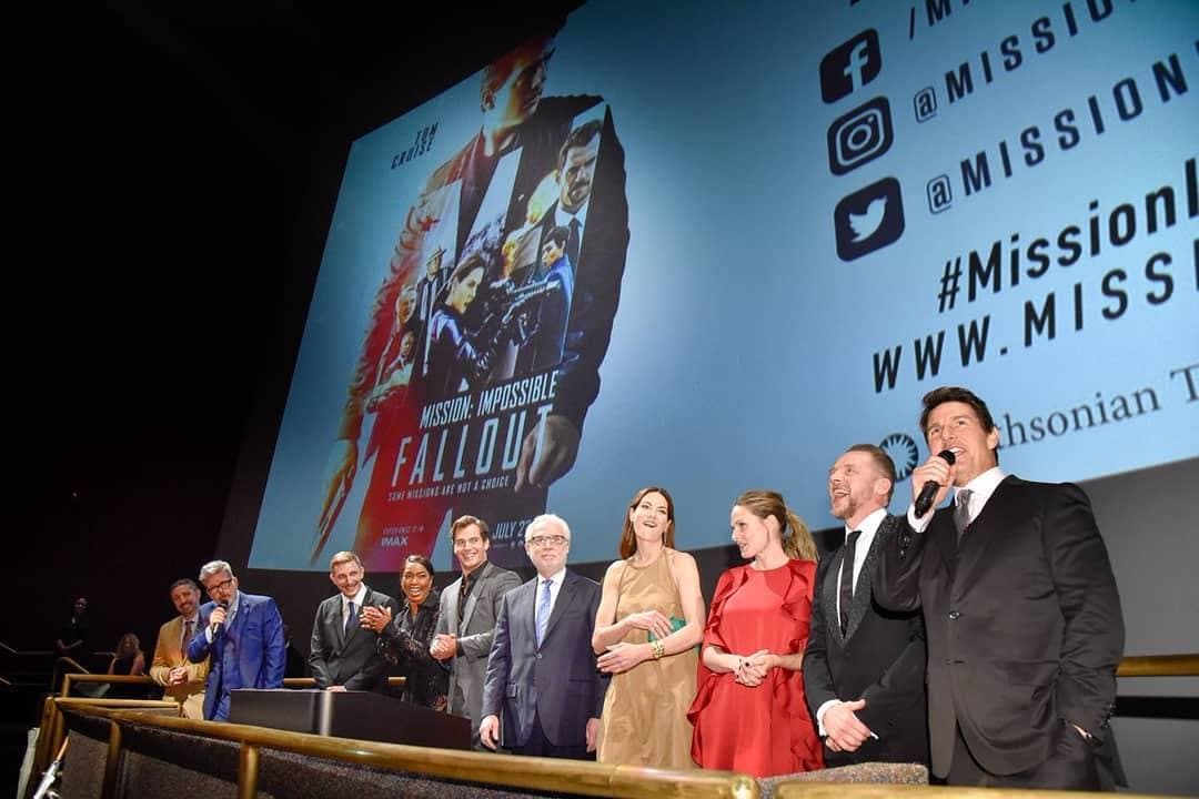A line of actors and the director in front of a cinema screen, Tom Cruise speaking into a microphone at the right