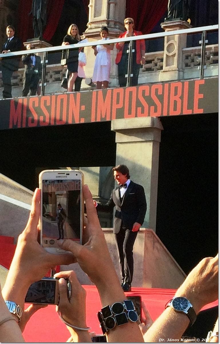Tom Cruise in a dark suit on a red carpet under a Mission: Impossible banner, fans holding up phones in the foreground