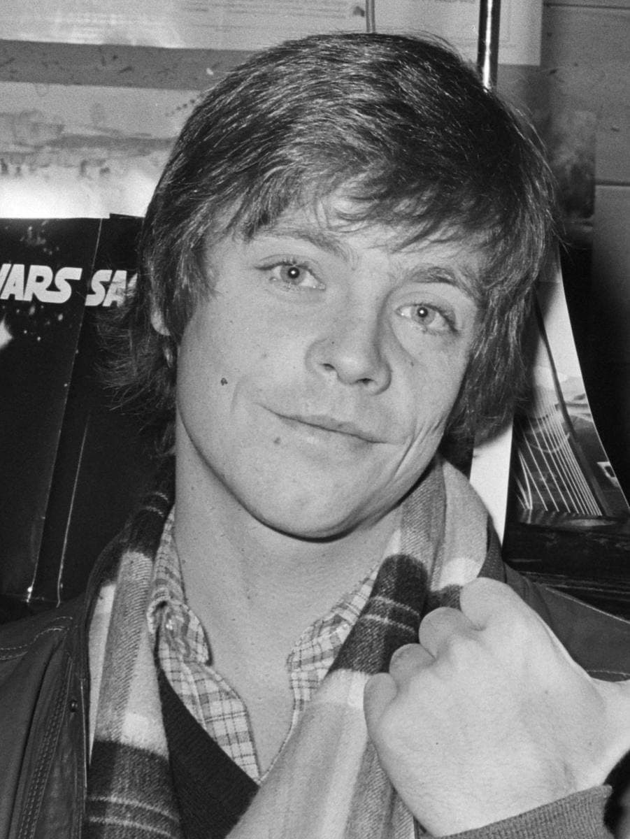 A young Mark Hamill with a tartan scarf, smiling, in black and white