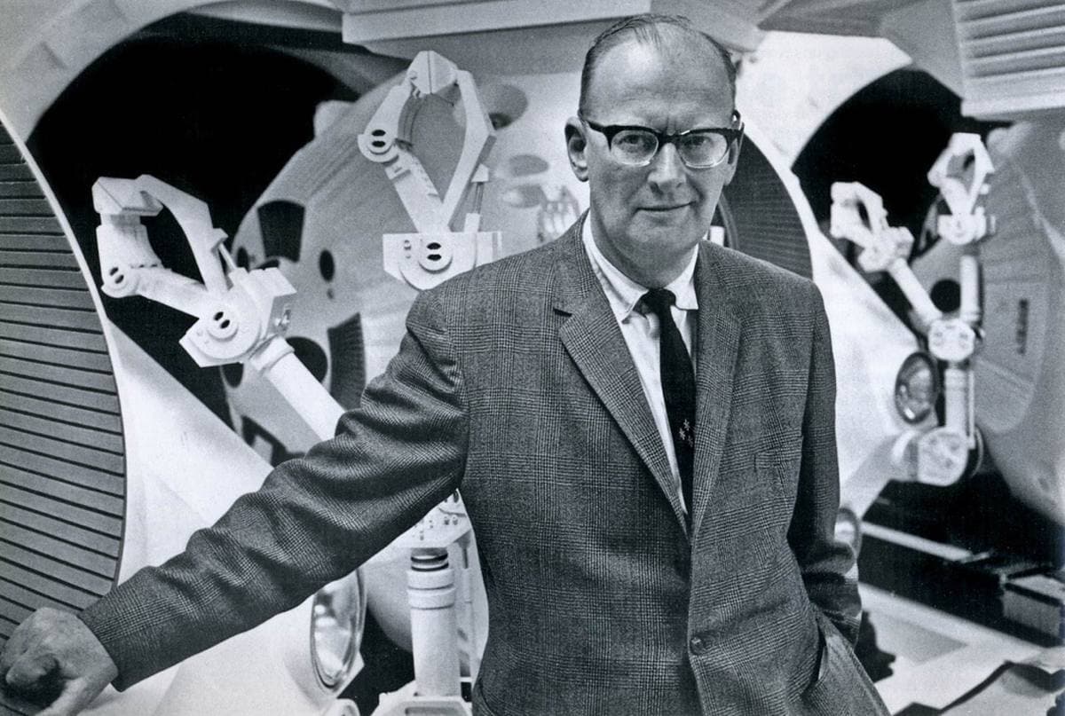 Arthur C. Clarke in a tweed jacket and glasses, leaning on a white spacecraft mock-up with robot arms behind him
