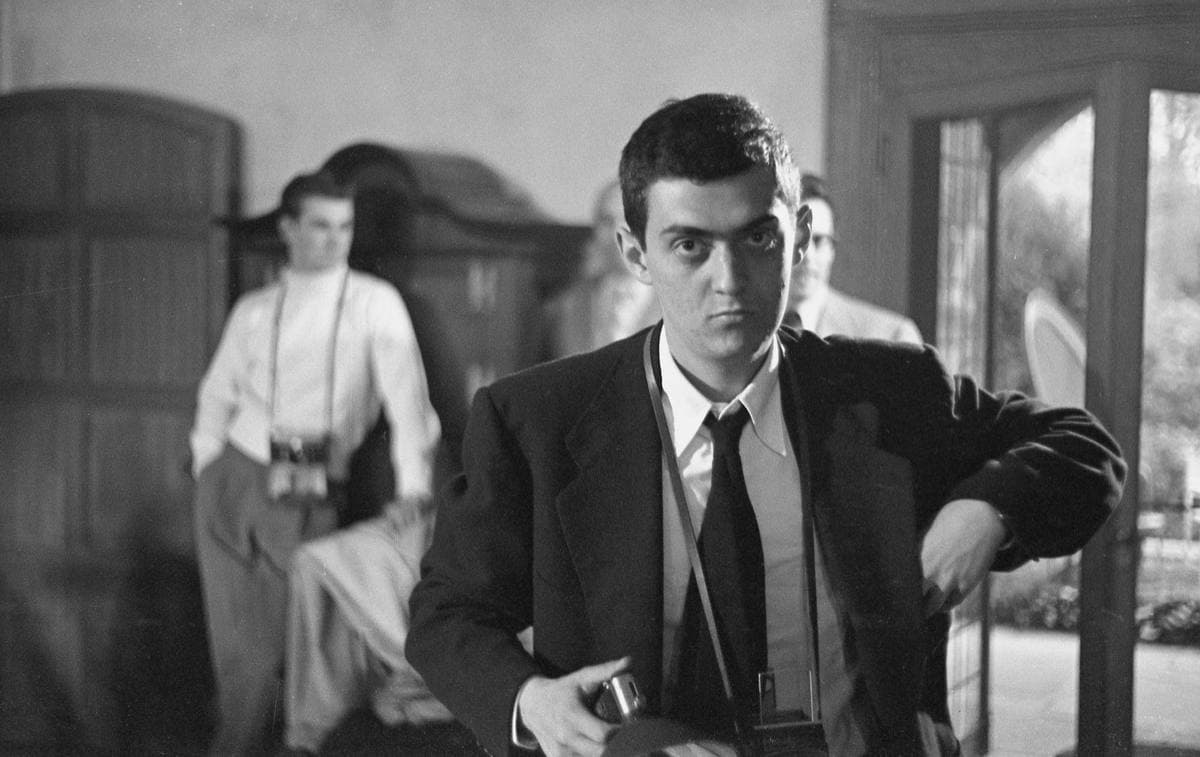 A young Stanley Kubrick in a dark jacket and tie, a camera in his hand, looking straight at the lens