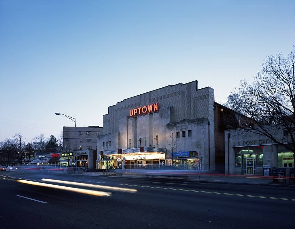 An art deco cinema with a red UPTOWN sign at dusk, car lights streaking past