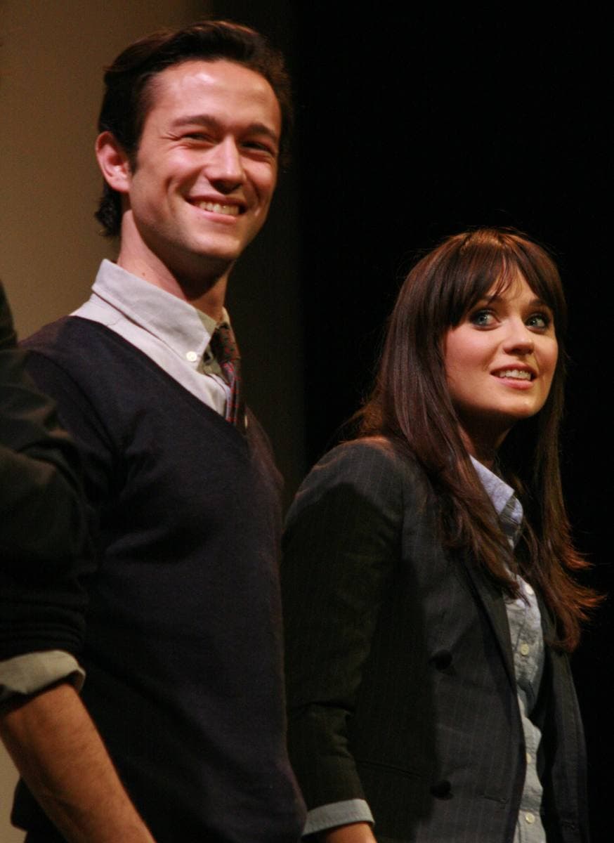 Joseph Gordon-Levitt, smiling in a dark jumper and tie, beside Zooey Deschanel with a fringe, against a black backdrop