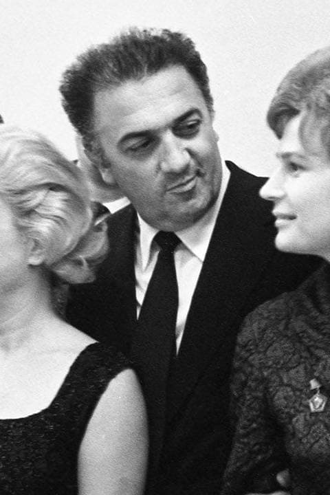 Federico Fellini in a dark suit and tie, smiling sideways