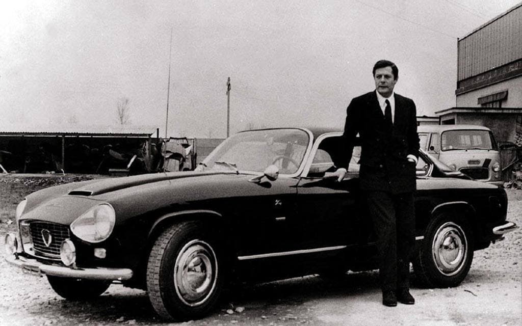 Marcello Mastroianni in a dark suit standing beside a black sports car