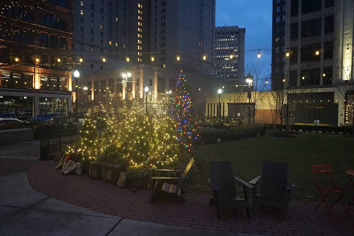 A small downtown square at dusk, its trees strung with Christmas lights, between tall buildings