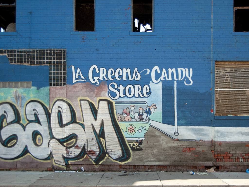 A blue brick wall on Chene Street painted with graffiti letters and a sign for La Greens Candy Store, with boarded windows above