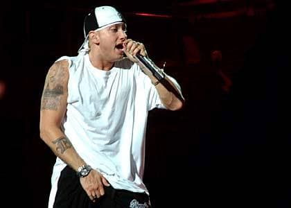Eminem in a white T-shirt and a backwards cap rapping into a microphone on a dark stage