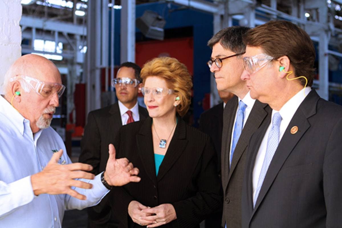 Visitors in safety glasses talking on the floor of a stamping plant, with steel press frames behind them