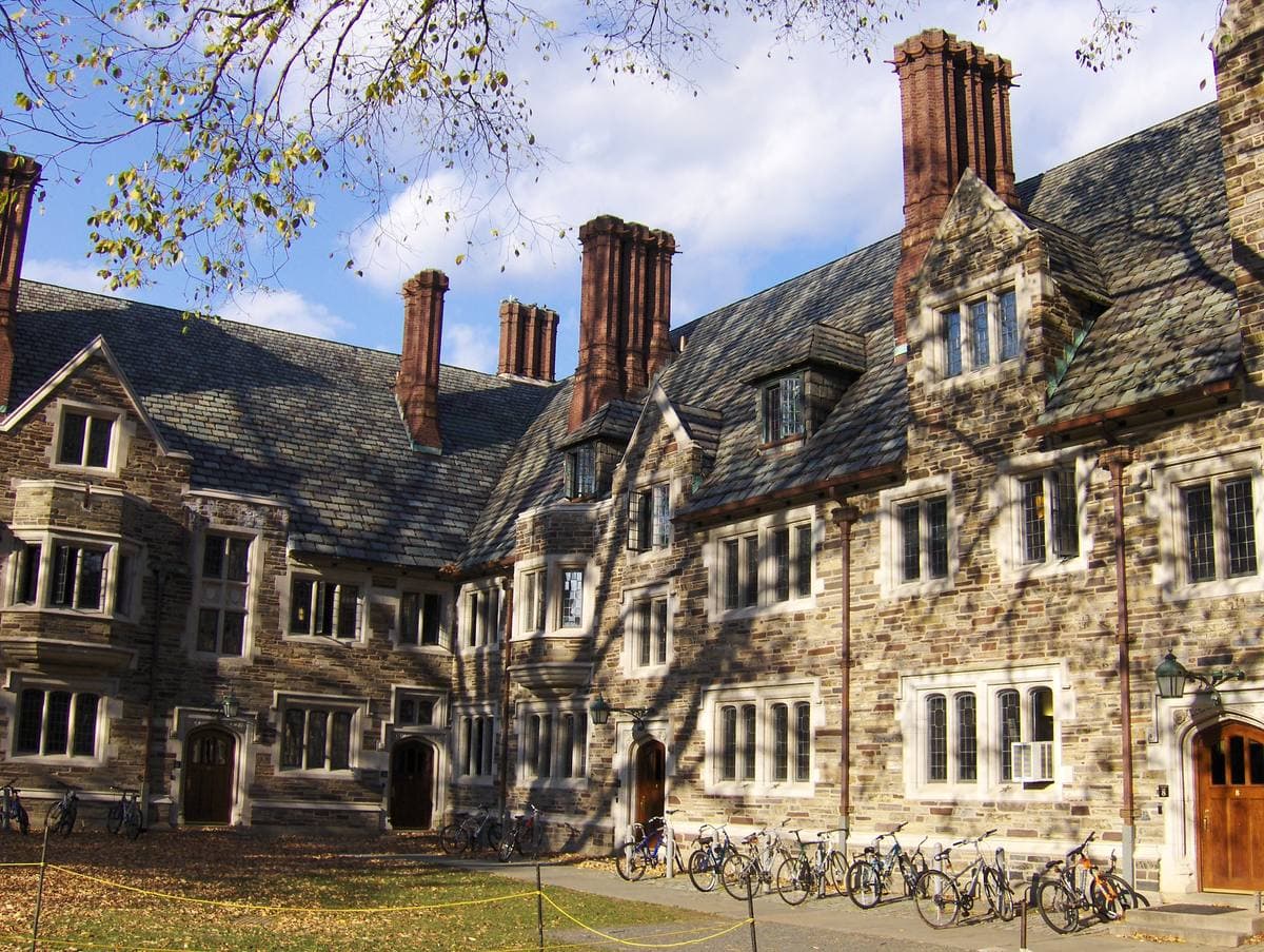 Stone Collegiate Gothic dormitory ranges with tall chimneys around a grassy courtyard, autumn trees, bicycles by a doorway