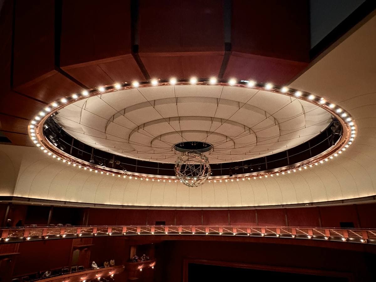 A round lit ceiling ring above the tiers of a red concert hall