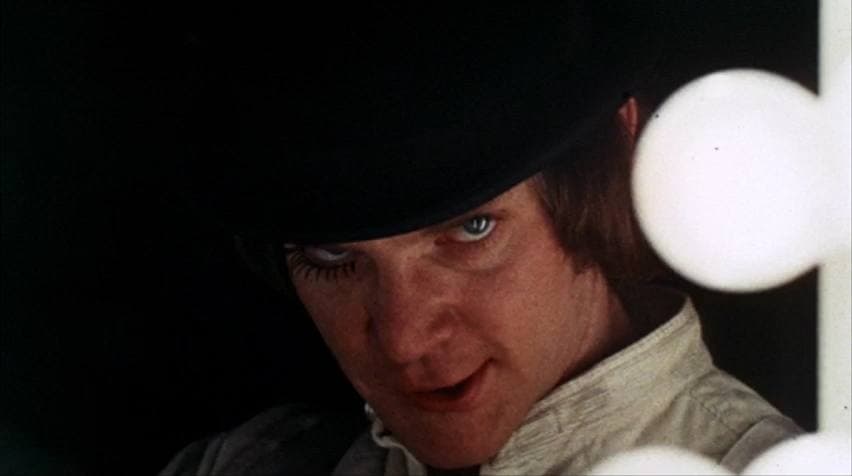 Malcolm McDowell as Alex in a bowler hat and false eyelashes, looking up with a half smile