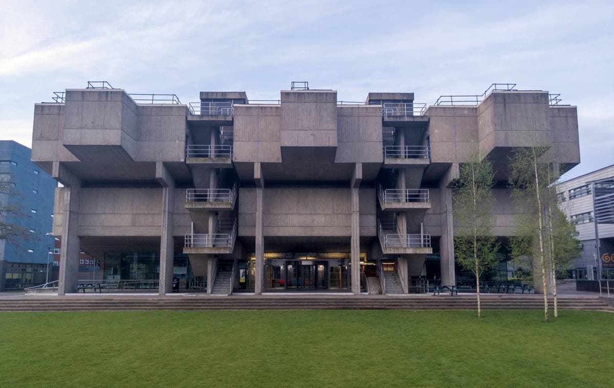 A grey concrete block of stacked, cantilevered lecture halls above a glazed ground floor and a lawn