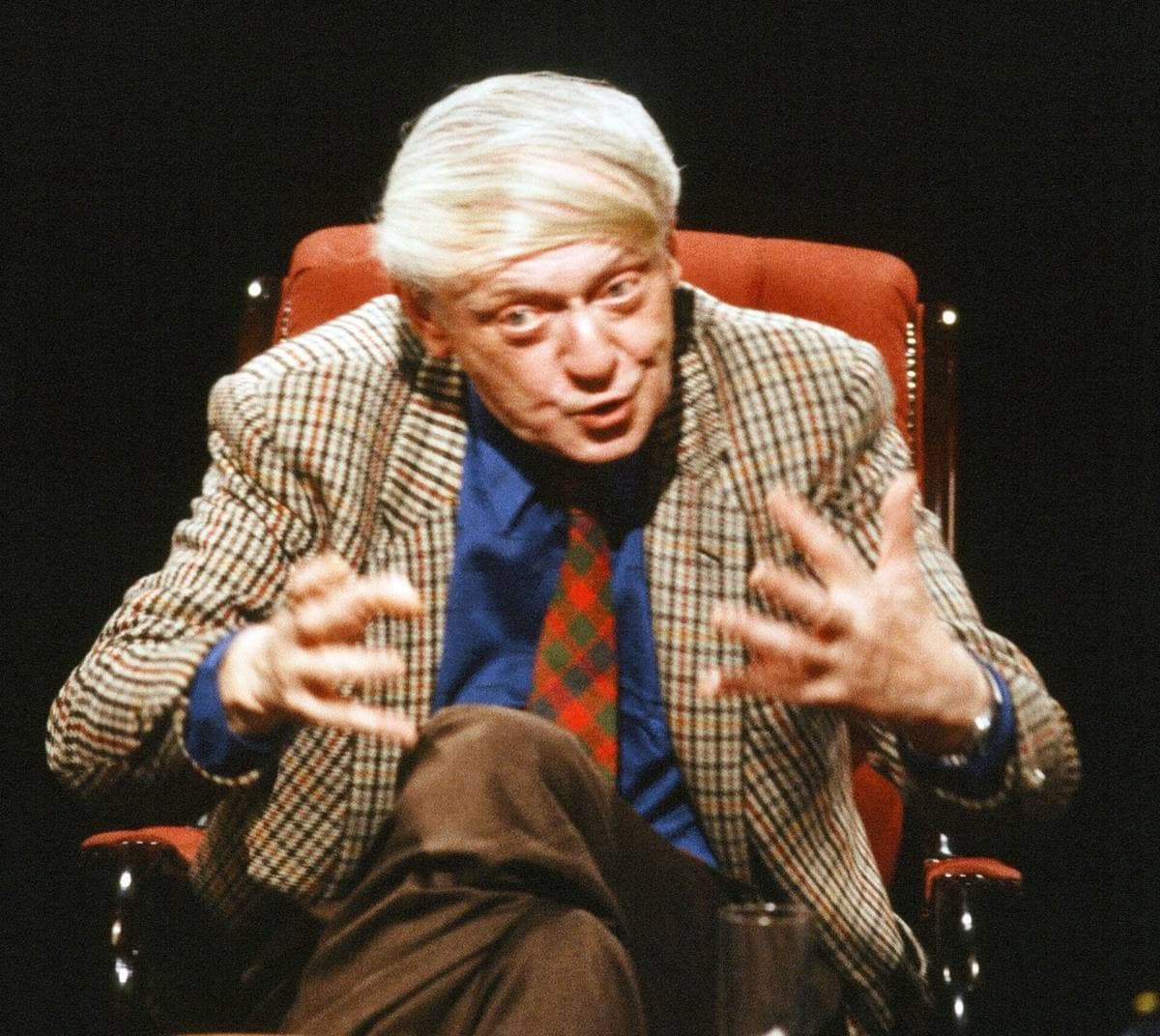 Anthony Burgess in a checked jacket and red tie, hands raised mid-sentence, in an armchair