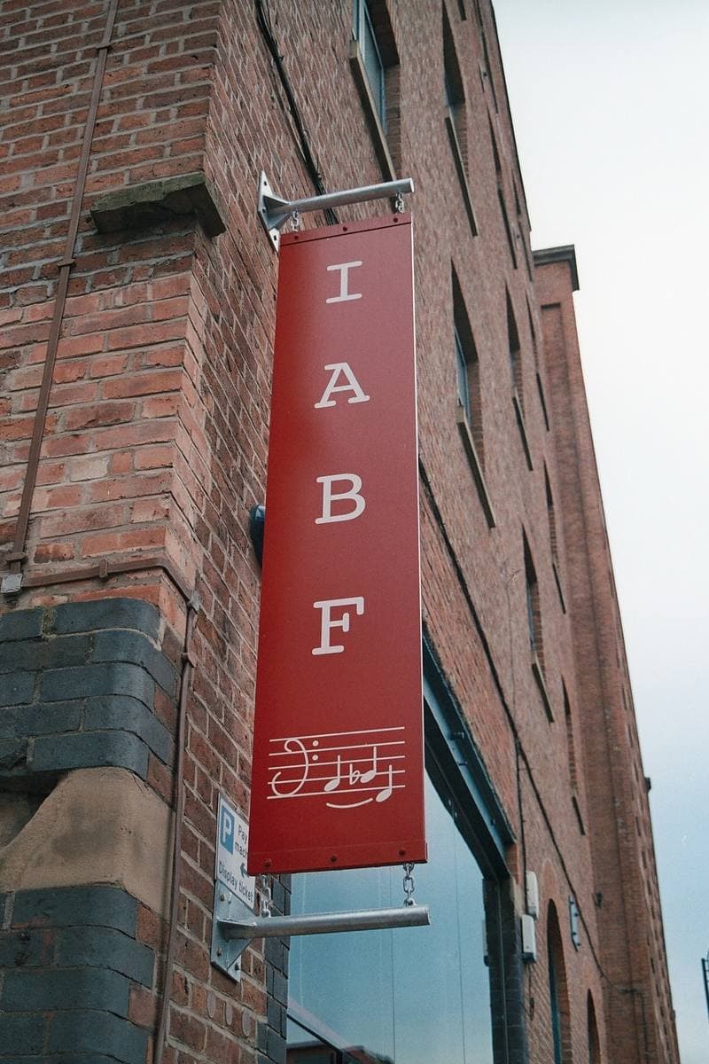 A tall red hanging sign reading I A B F above a stave of music, on a brick mill