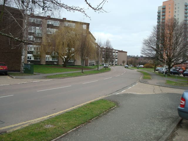 A curving suburban road with low brick flats and a tall tower block behind bare trees