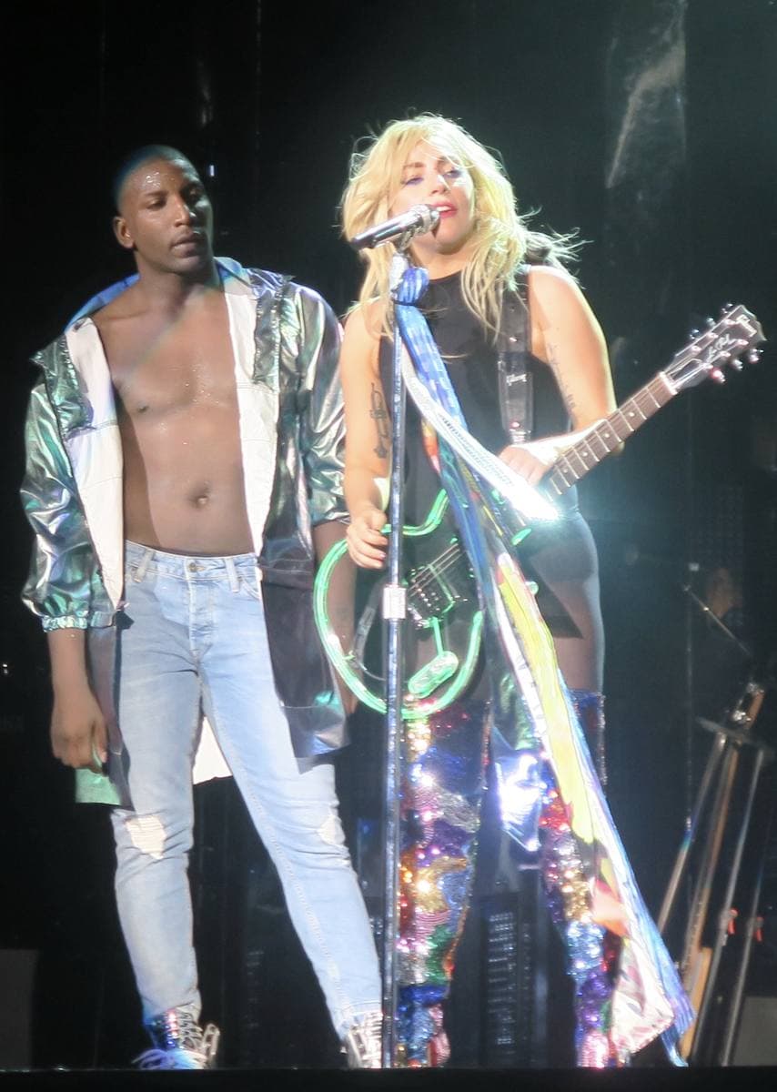 Lady Gaga on a festival stage with a guitar and a microphone, in a fringed jacket