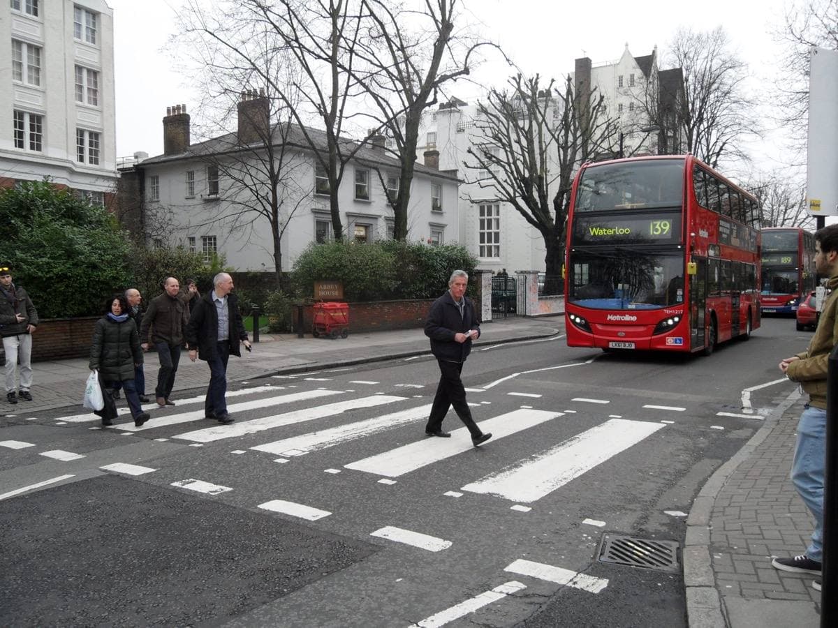 People walking over a zebra crossing on a leafy road, a red bus behind