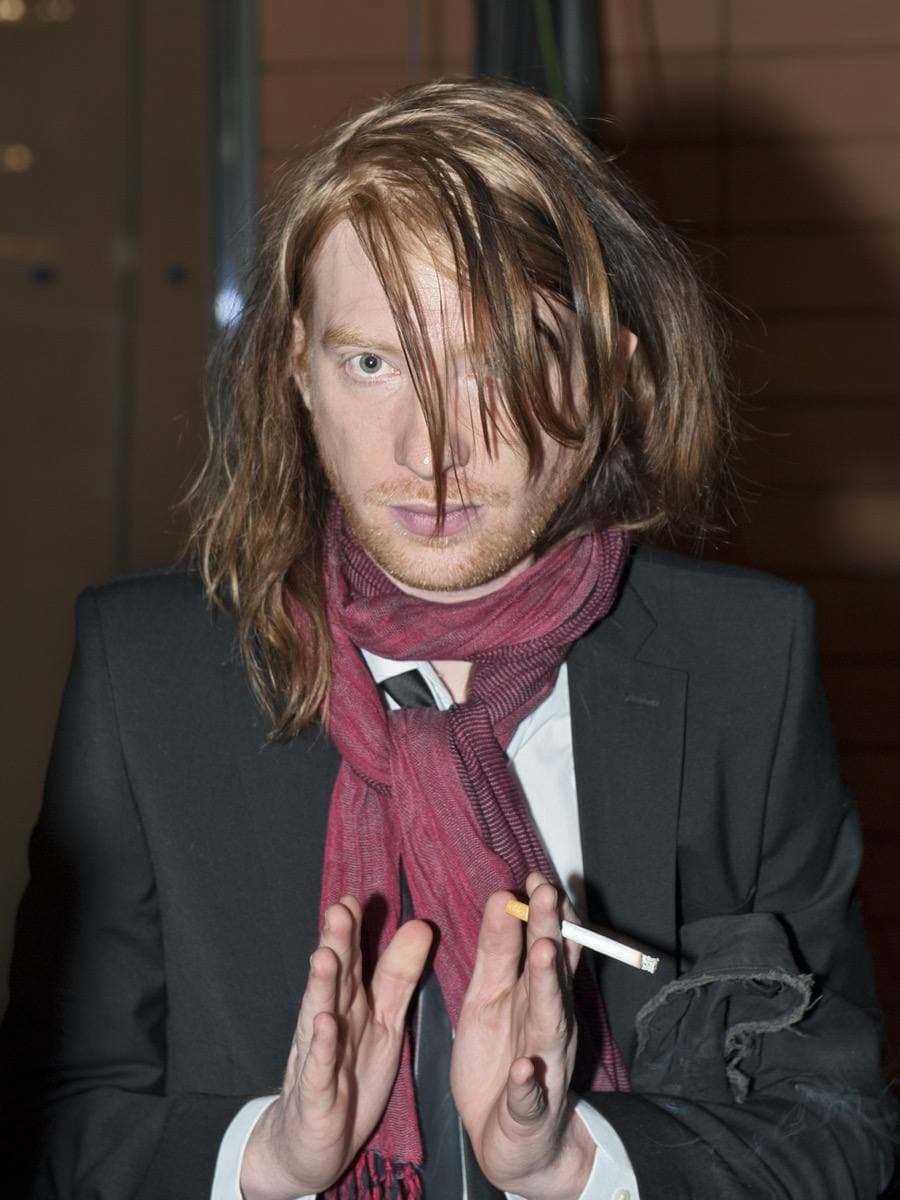 Domhnall Gleeson with long red hair and a dark suit, holding an award