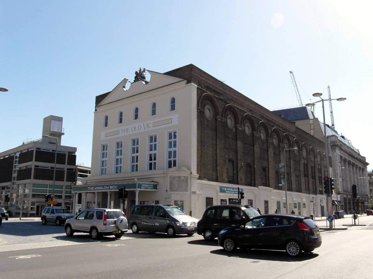 A pale classical theatre front on a street corner, cars outside
