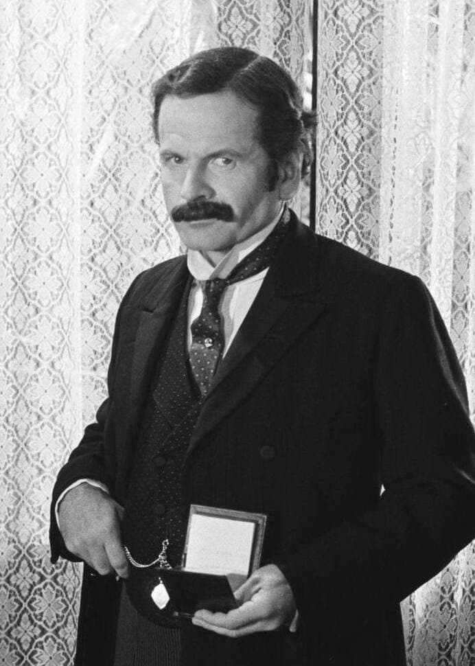 Ian Holm with a dark moustache in a Victorian frock coat, holding an open watch case, black and white