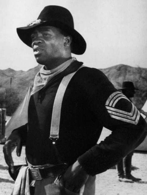 Yaphet Kotto in a wide-brimmed hat and cavalry jacket, speaking, black and white