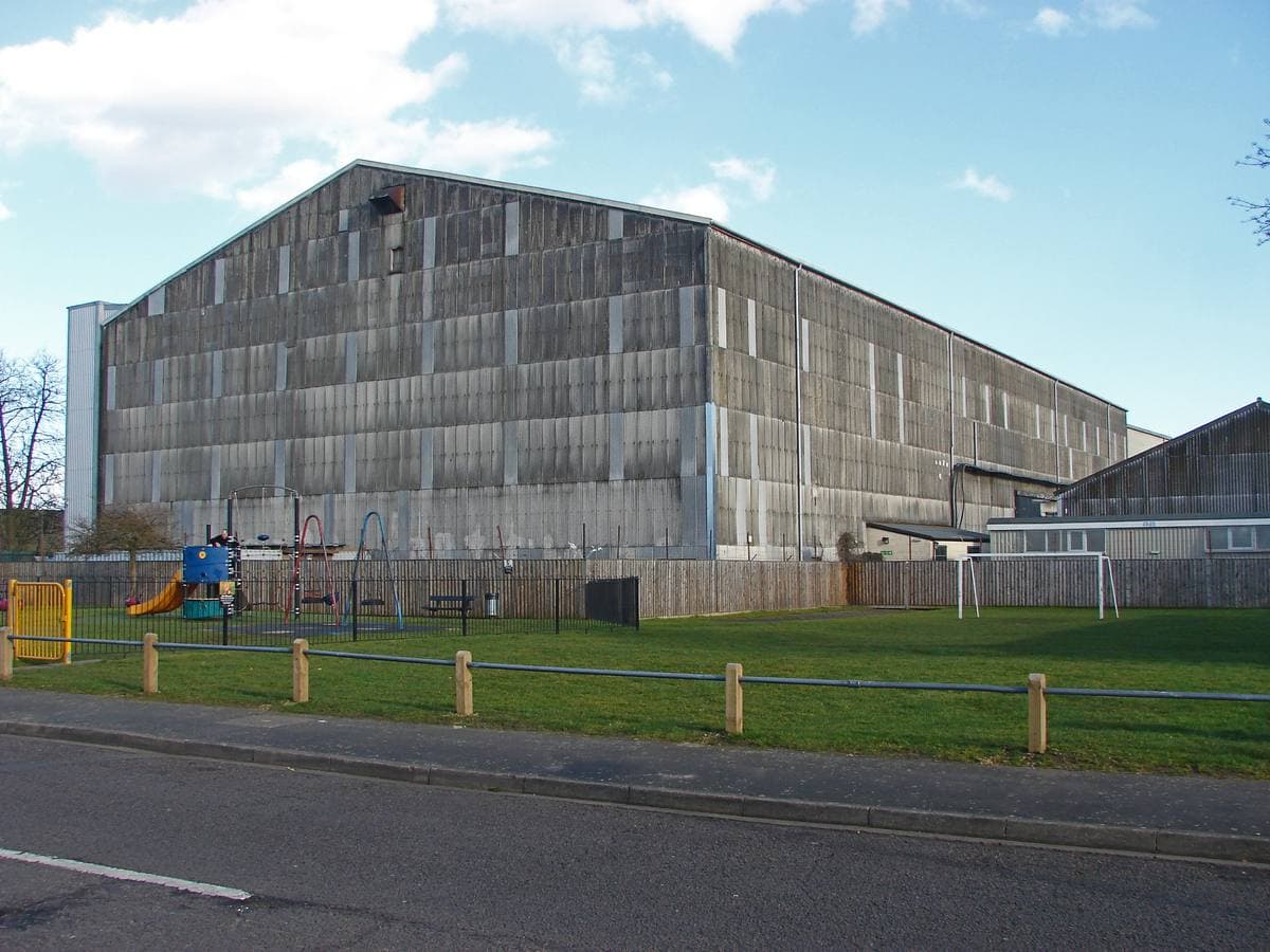 A huge corrugated grey stage building behind a green verge and a low fence