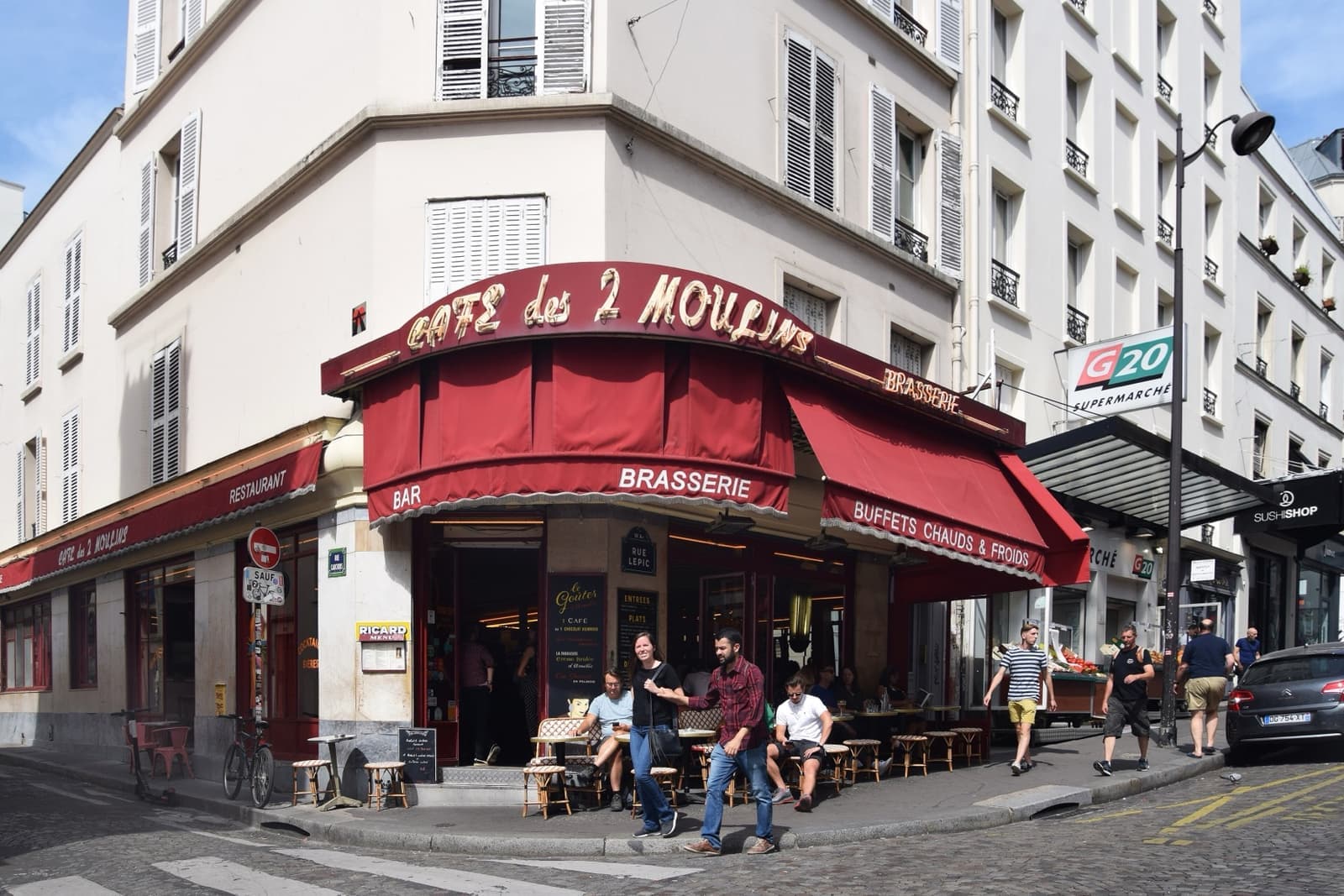 The Café des 2 Moulins on a street corner, with a red awning and tables on the pavement