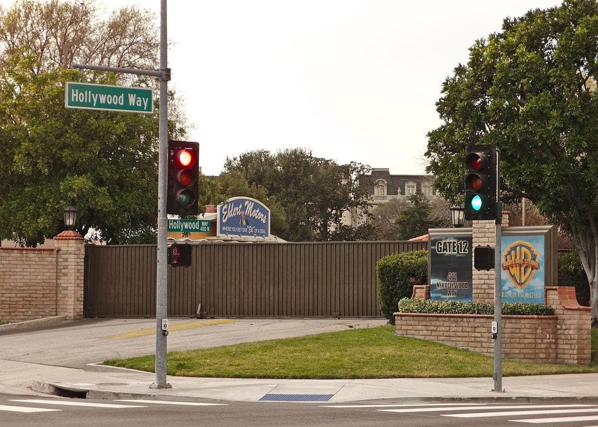 A studio gate with a Warner Bros. sign behind a brick wall at the corner of Hollywood Way