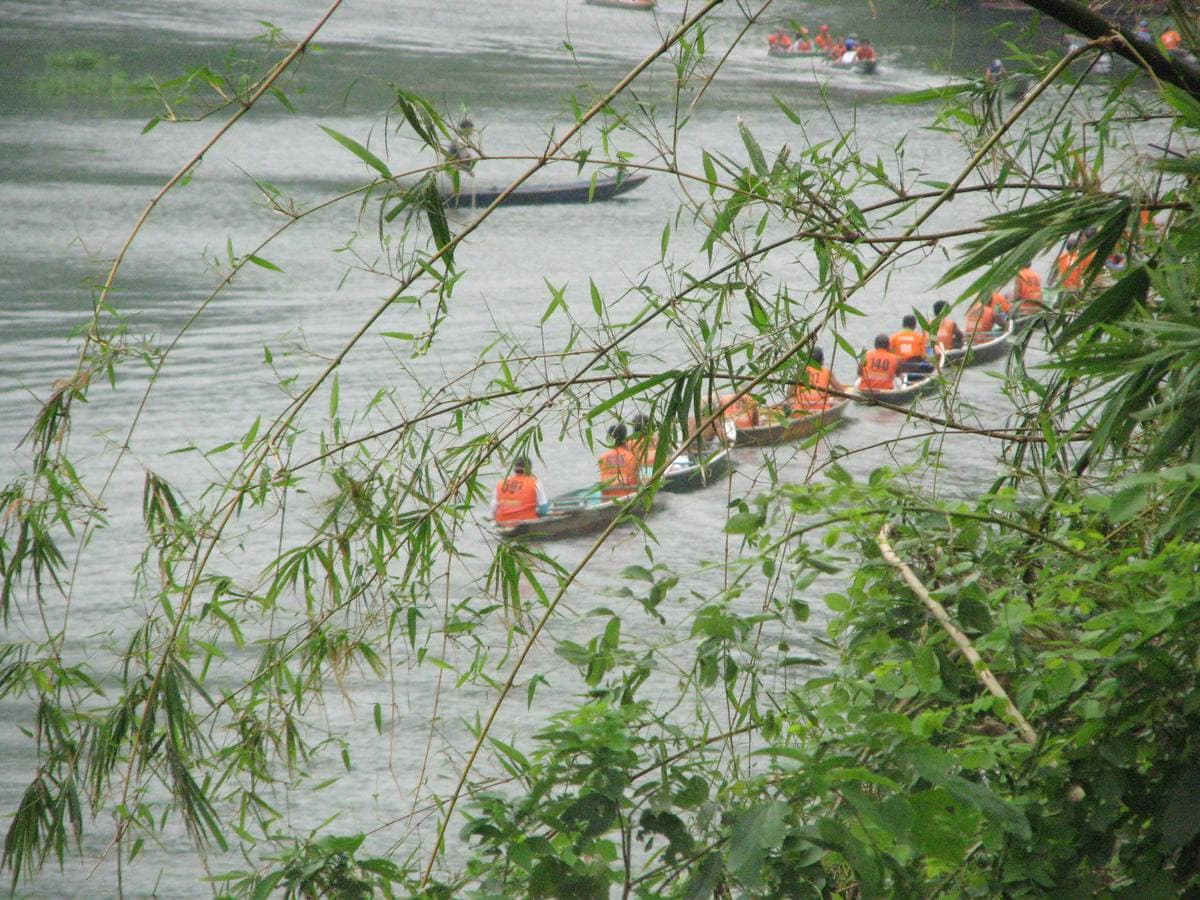 A line of narrow wooden boats with passengers on a brown river under jungle cliffs