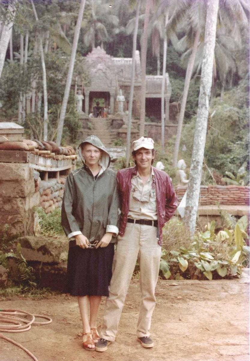 A woman in a hooded jacket and a man in a cap stand on a red-earth clearing in front of a stone temple set among palm trees