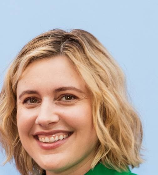 Greta Gerwig smiling, shoulder-length wavy fair hair, against a pale blue wall
