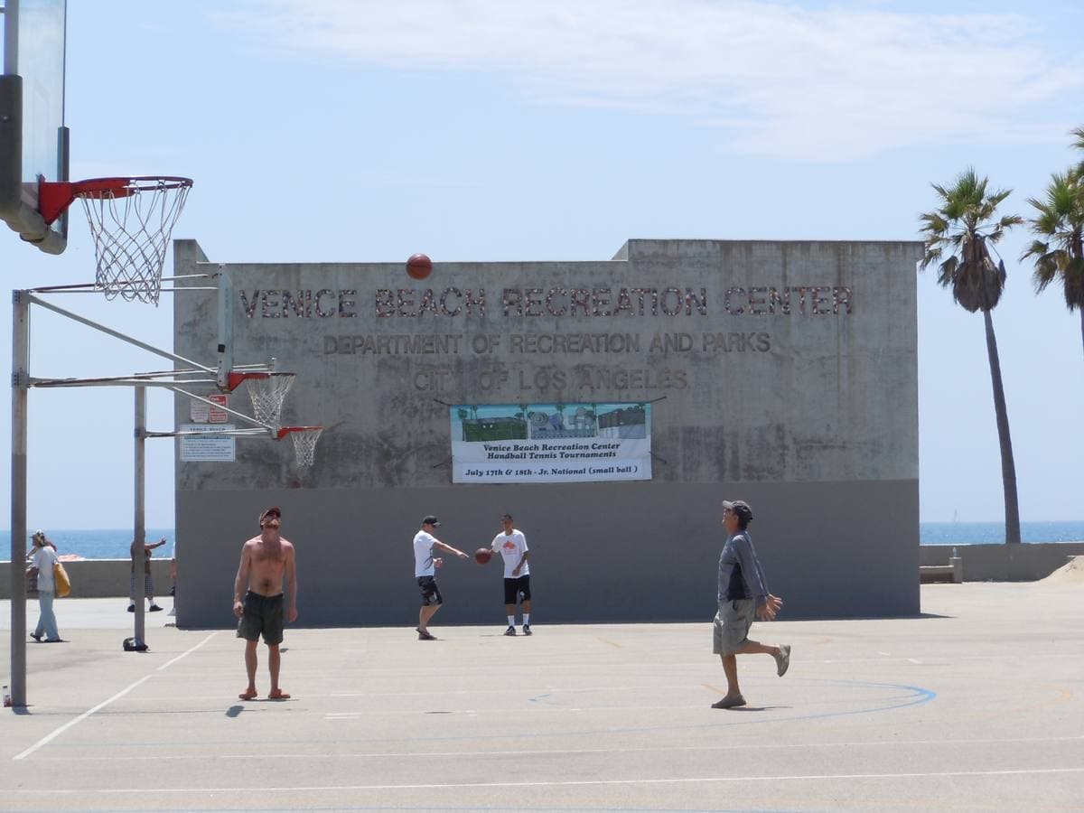 Men playing basketball on an outdoor court in front of a low concrete building with palms behind
