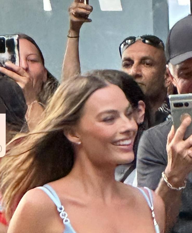 Margot Robbie smiling, hair blowing, in a pale blue satin dress, with photographers behind her