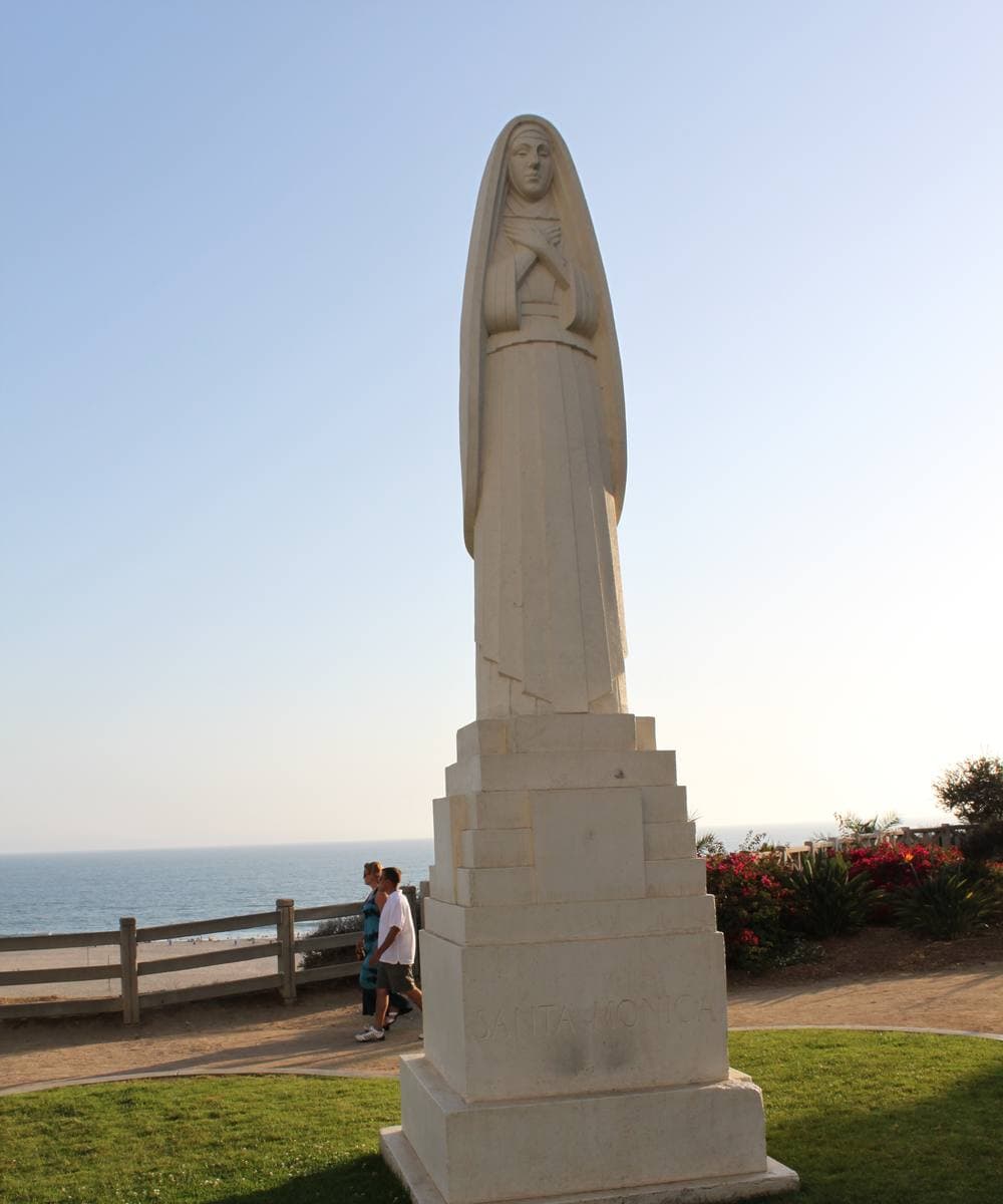 A tall pale stone statue of a robed woman on a plinth, ocean behind