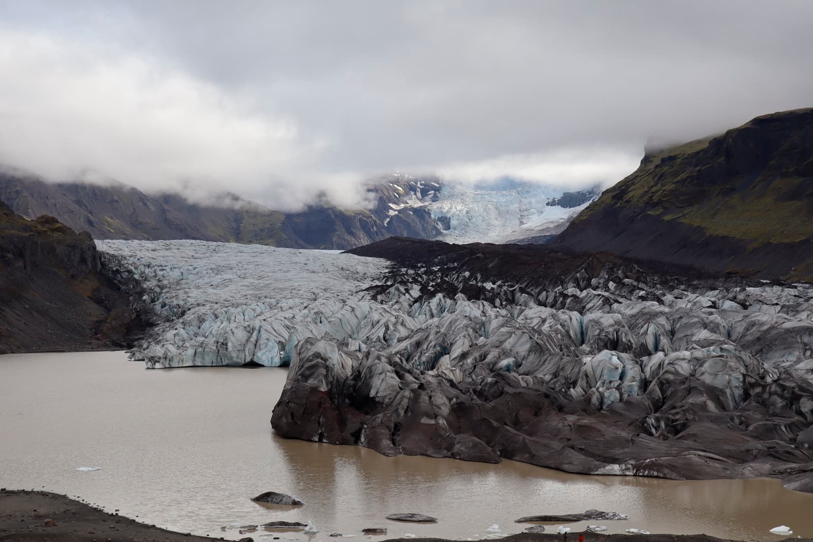 A grey, crevassed glacier tongue pours between dark mountain walls into a meltwater lake under low cloud