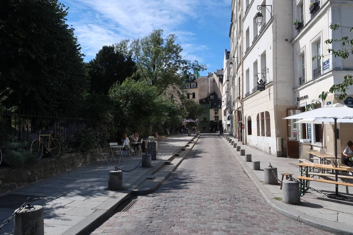A short cobbled street with trees on one side and pale houses on the other