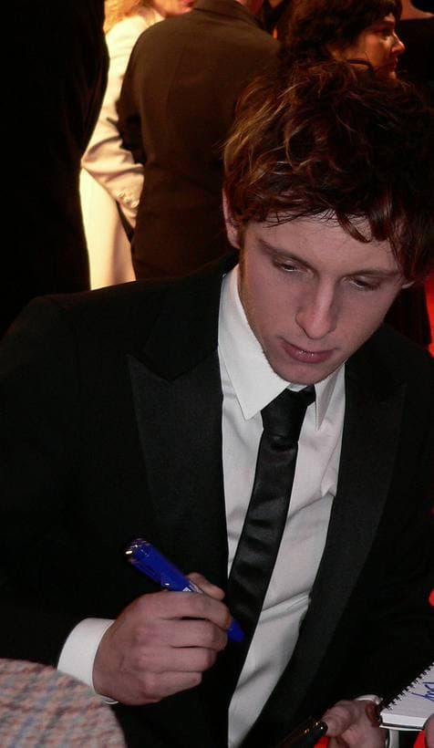 Colour photograph of a young man with tousled brown hair in a dark suit, signing autographs on a red carpet