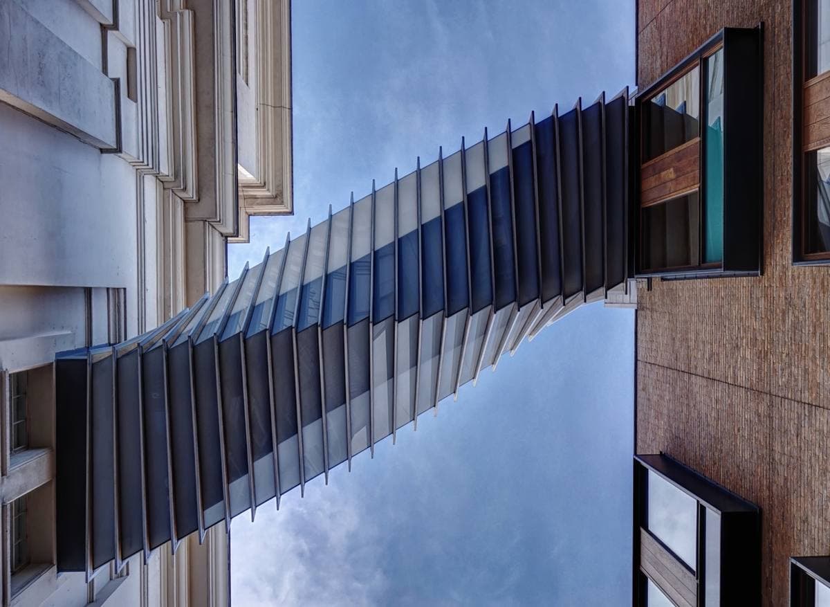 A twisting glass and aluminium bridge seen from below between two buildings