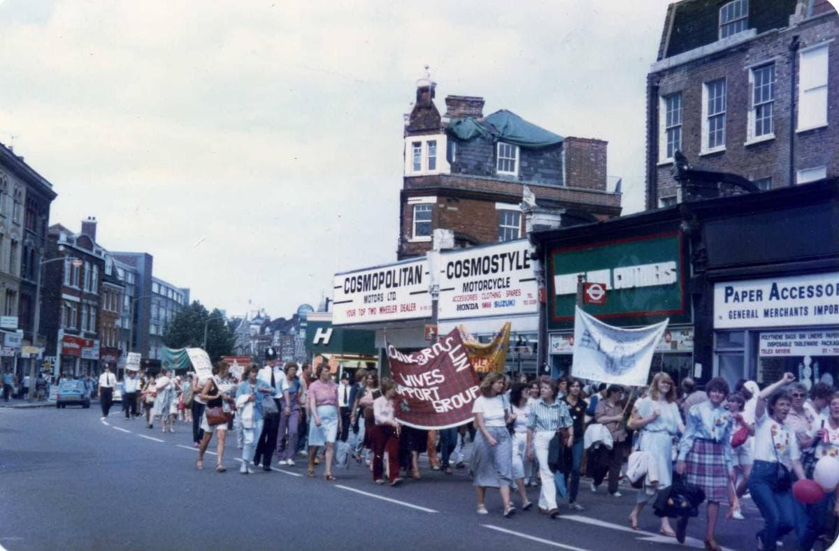 Colour photograph of a crowd of women marching down a high street behind hand-made banners, shopfronts behind