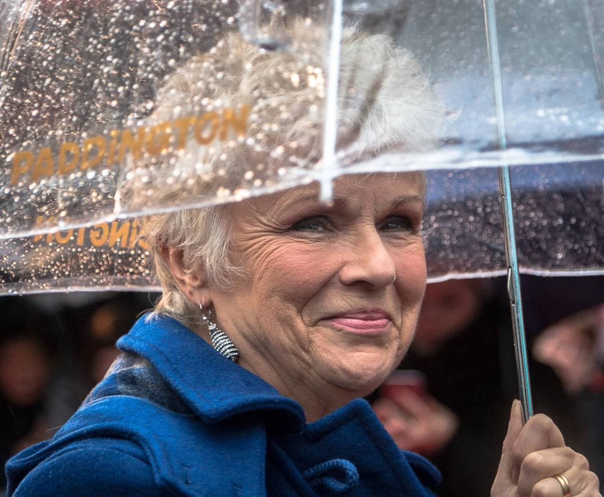 Julie Walters smiling under a clear umbrella in the rain